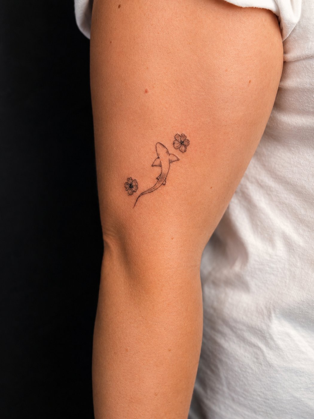 Fine-line tattoo of a small shark framed by two delicate flowers on an upper arm